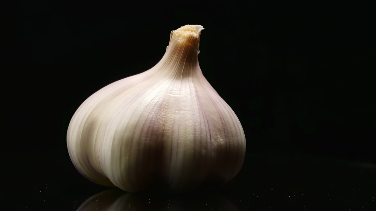 Bulbo d'aglio non sbucciato che poggia su una superficie nera riflettente su uno sfondo nero profondo, catturato in una vista ravvicinata dal basso con luce soffusa, forte contrasto, texture della buccia dettagliata e isolamento pulito
