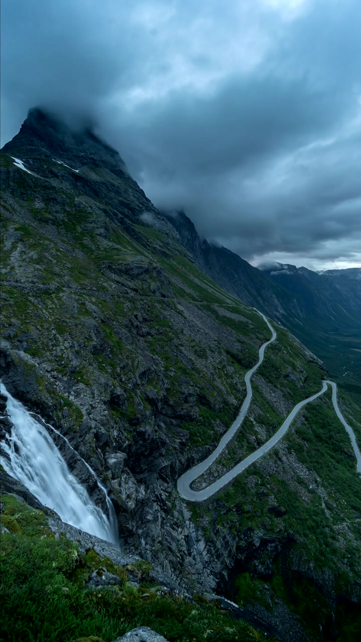 Vista verticale di cascate che scorrono attraverso la montagna con Trollstigen, strada di montagna serpeggiante a Rauma, More og Romsdal, Norvegia. - ripresa in timelapse