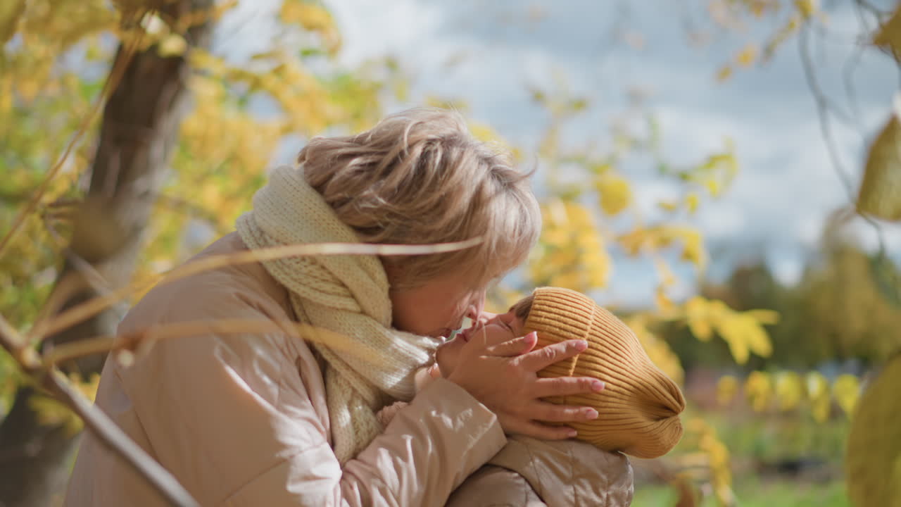 mother tenderly kisses daughter wearing mustard beanie and jacket amid golden autumn leaves gently moving in breeze, sharing warm emotional connection during calm day