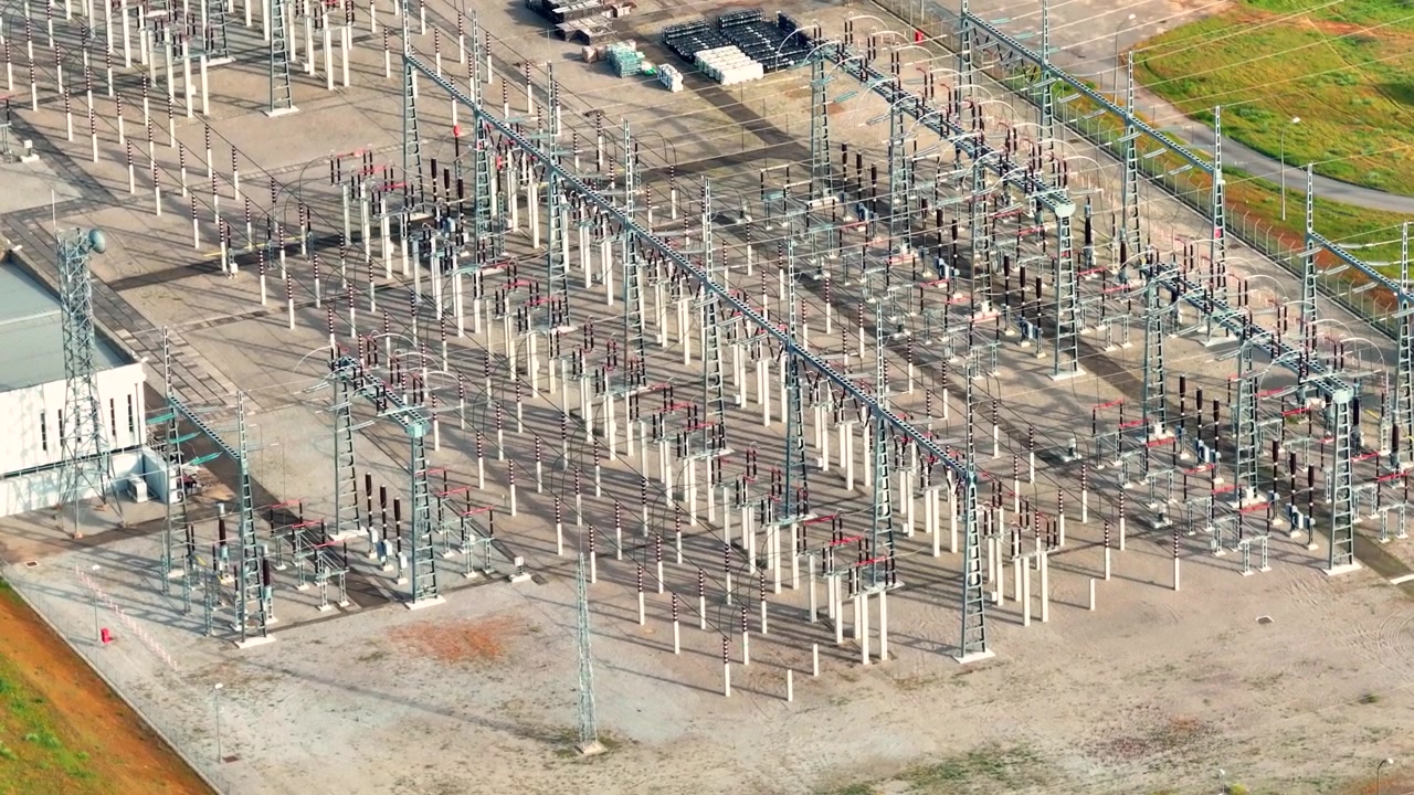 Aerial view of power substation showing electrical equipment and transmission lines. Structures support energy distribution within electrical grid. Nearby vehicles indicate maintenance activity.