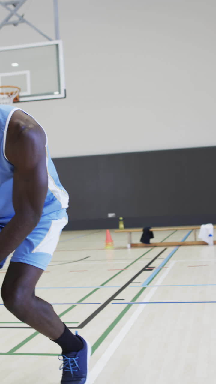 Vertical video of male african american basketball player training in indoor court, in slow motion