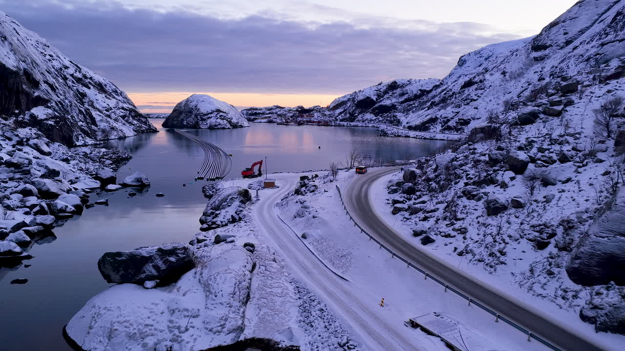 Atemberaubende Drohnenaufnahme der schneebedeckten Lofoten-Inseln in Norwegen während der Dämmerung, mit einer kurvenreichen Straße und ruhigem Fjordwasser.