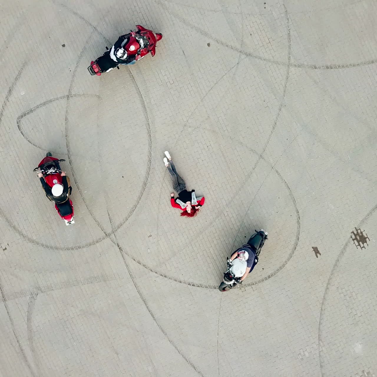 Three professional bikers on their motorcycles riding on a road in the form of a circle. Smiling beautiful young woman lying in the centre of the pavement. Camera moves top down.