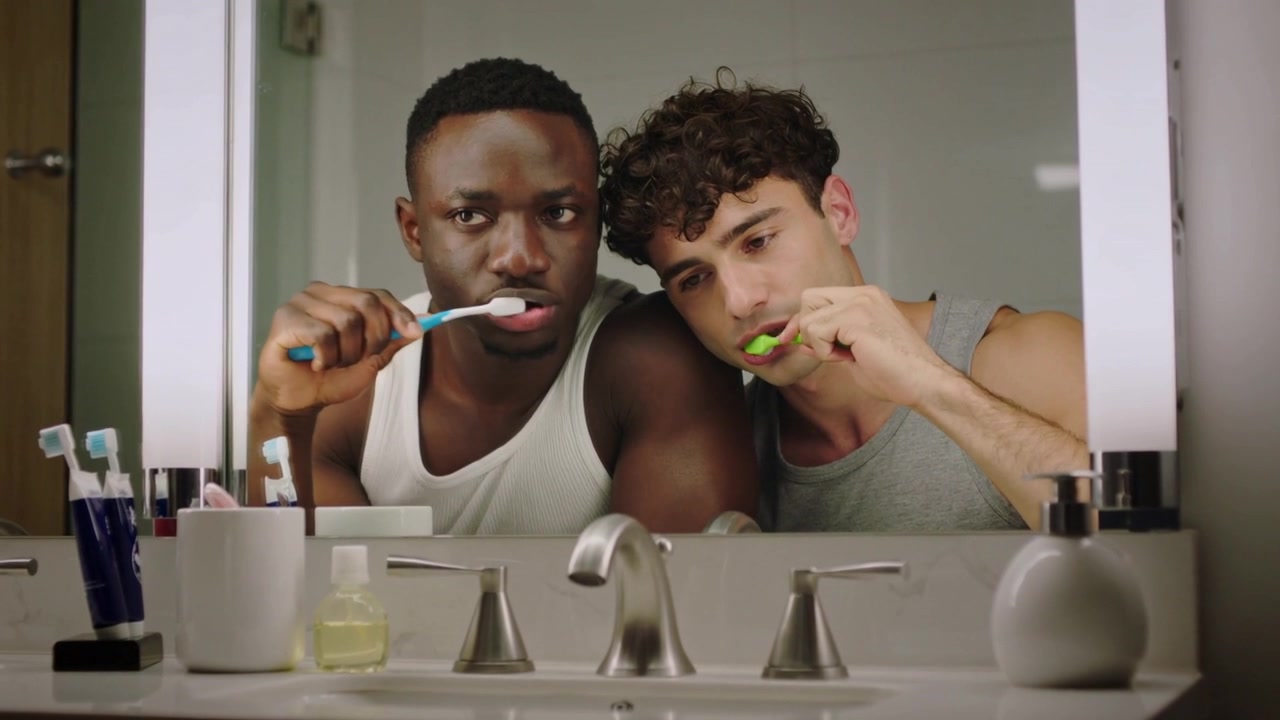 Waking, two men white and gray tops brushing teeth at mirror holding blue green brushes preparing