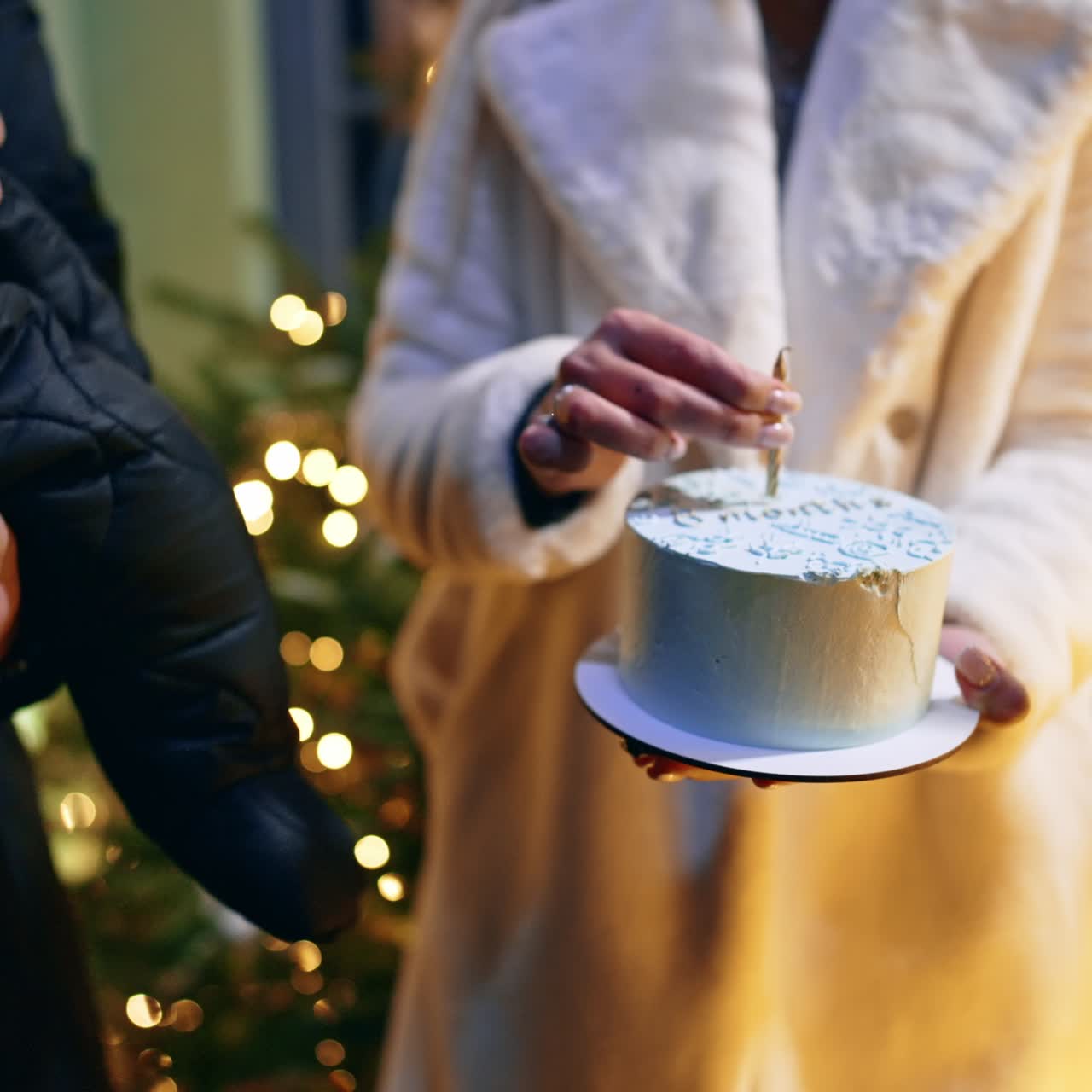 Donna irriconoscibile in camice bianco tiene una torta bianca con una candela sopra. Un uomo con in braccio un bambino è in piedi accanto. Primo piano