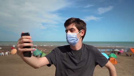 young man in face mask taking a selfie showing peace sign at the beach at summertime