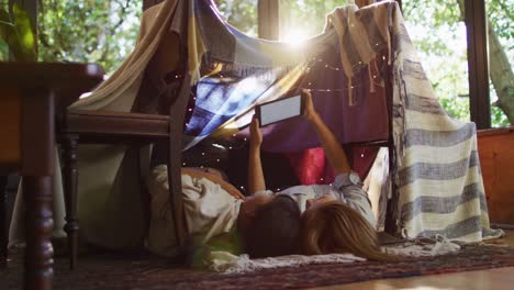 Asian-mother-and-daughter-using-digital-tablet-while-lying-under-blanket-fort-at-home