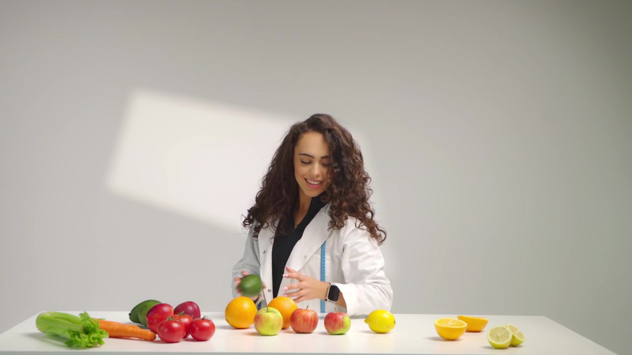 Nutricionista feminina sorridente segura frutas e vegetais frescos enquanto olha para a câmera em um cenário de estúdio cinematográfico brilhante. Conceito de alimentação saudável, planejamento de dieta e bem-estar em câmera lenta