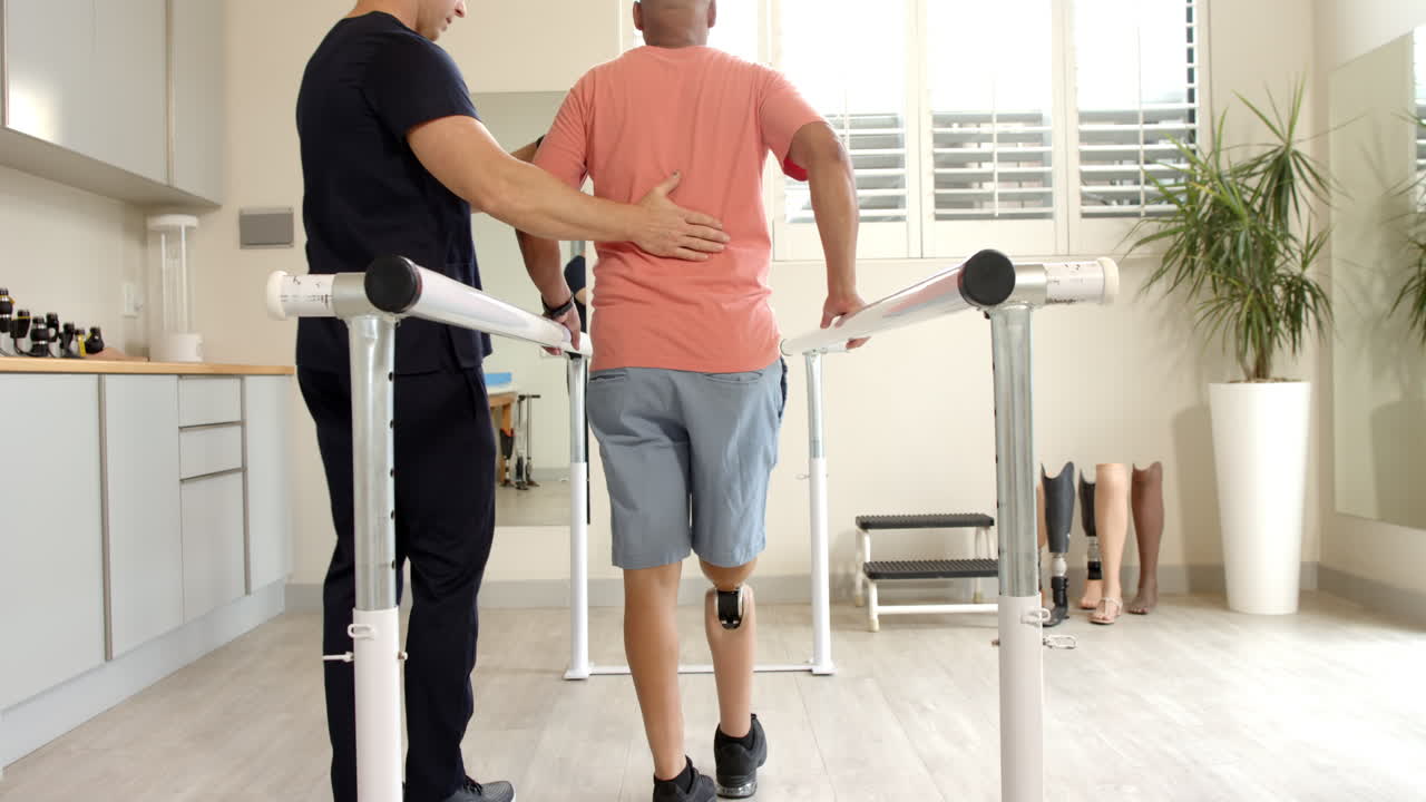 Therapist assisting man with prosthetic leg walking on parallel bars ...