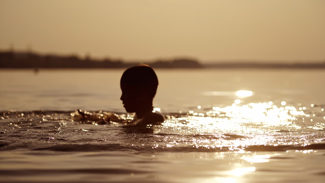 Young boy plays with water in the evening river. Silhouette of a child making waves on natural water background at sunset. Healthy lifestyle.