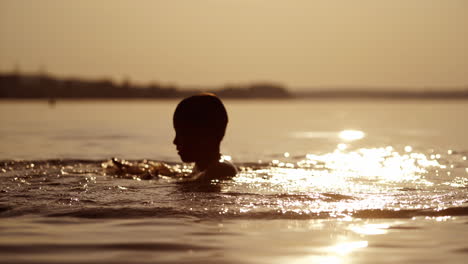 young boy plays with water in the evening river. silhouette of a child making waves on natural water background at sunset. healthy lifestyle.