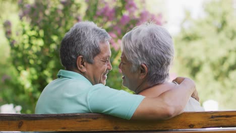 Video-of-happy-biracial-senior-couple-embracing-and-sitting-on-bench-in-garden