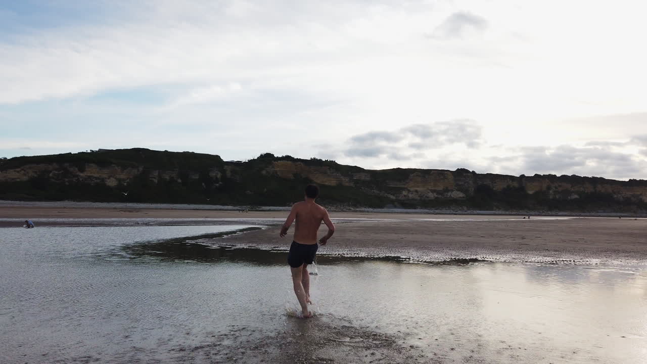 A shirtless man sprinting across wet sand on an Omaha beach to retrieve a frisbee. A moment of energy, fun, and playful pursuit in a vast coastal landscape.