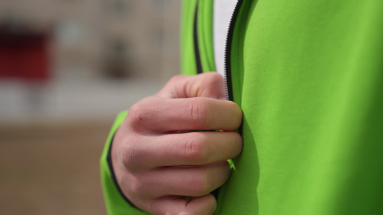 cremallera de chaqueta, imagen detallada que muestra una mano abrochando la cremallera de una chaqueta verde durante un trayecto urbano, primer plano que captura una mano ajustando la cremallera de una prenda exterior verde antes de un viaje por la ciudad