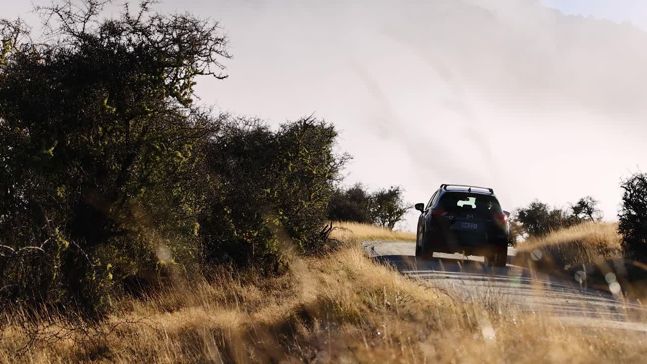 A dark SUV travels on a winding rural road through dry grassland and sparse trees, under soft natural daylight, with a steady rear-facing camera angle