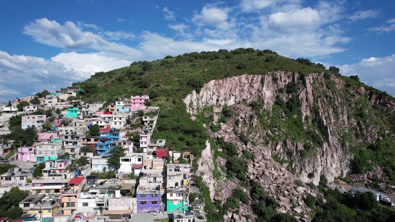 Drone, Ecatepec'teki Cerro Gordo'ya doğru uçarak parlak mavi gökyüzü ve dramatik bulutların altında kayalık uçurumu, renkli yamaç evlerini ve yemyeşil bitki örtüsünü ortaya çıkarıyor