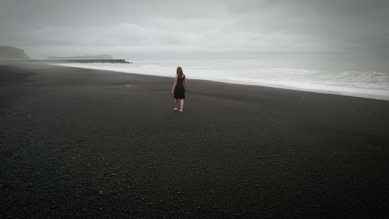 una joven hermosa con un vestido negro caminando por la playa de arena negra de islandia, dramáticas olas aéreas del océano