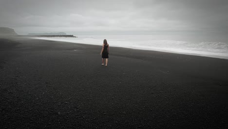 Young-beautiful-woman-in-black-dress-walking-on-black-sand-beach-Iceland,-dramatic-aerial-ocean-waves