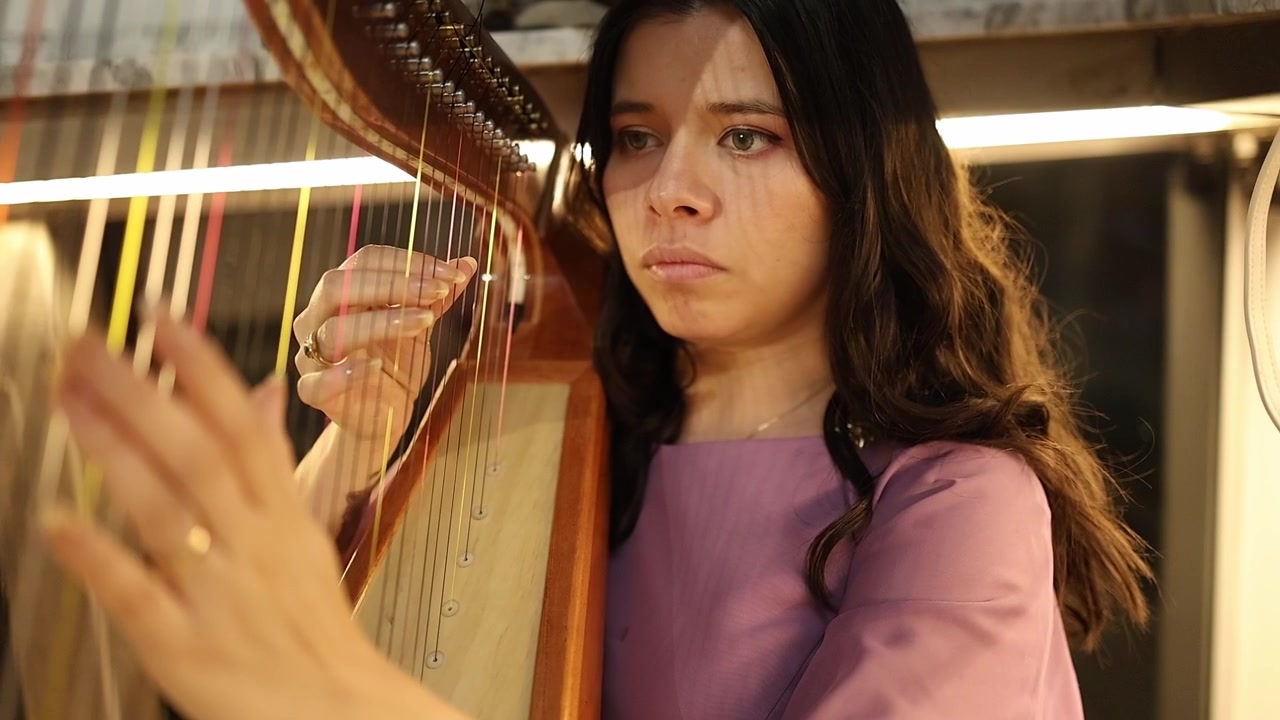 A focused young woman plays a harp in a warmly lit indoor setting. Shot in close-up, the camera captures expressive hand movements and emotional concentration for artistic music footage.