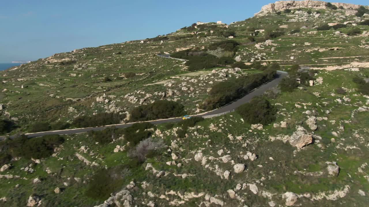 Colourful Car Driving Fast On The Road Up To The Lush Rocky Hill Under The Clear Blue Sky - Aerial Shot