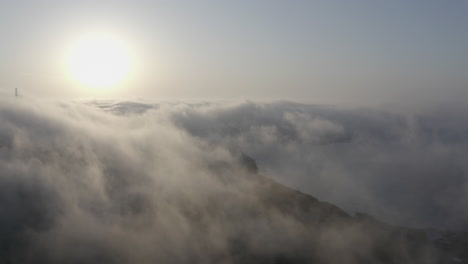 Epische-Vogelperspektive-Auf-Die-Meeresküste-Bei-Sonnenuntergang-Mit-Dichten-Nebelwolken,-Die-über-Die-Hügel-Ziehen-Und-Sie-Vollständig-Bedecken