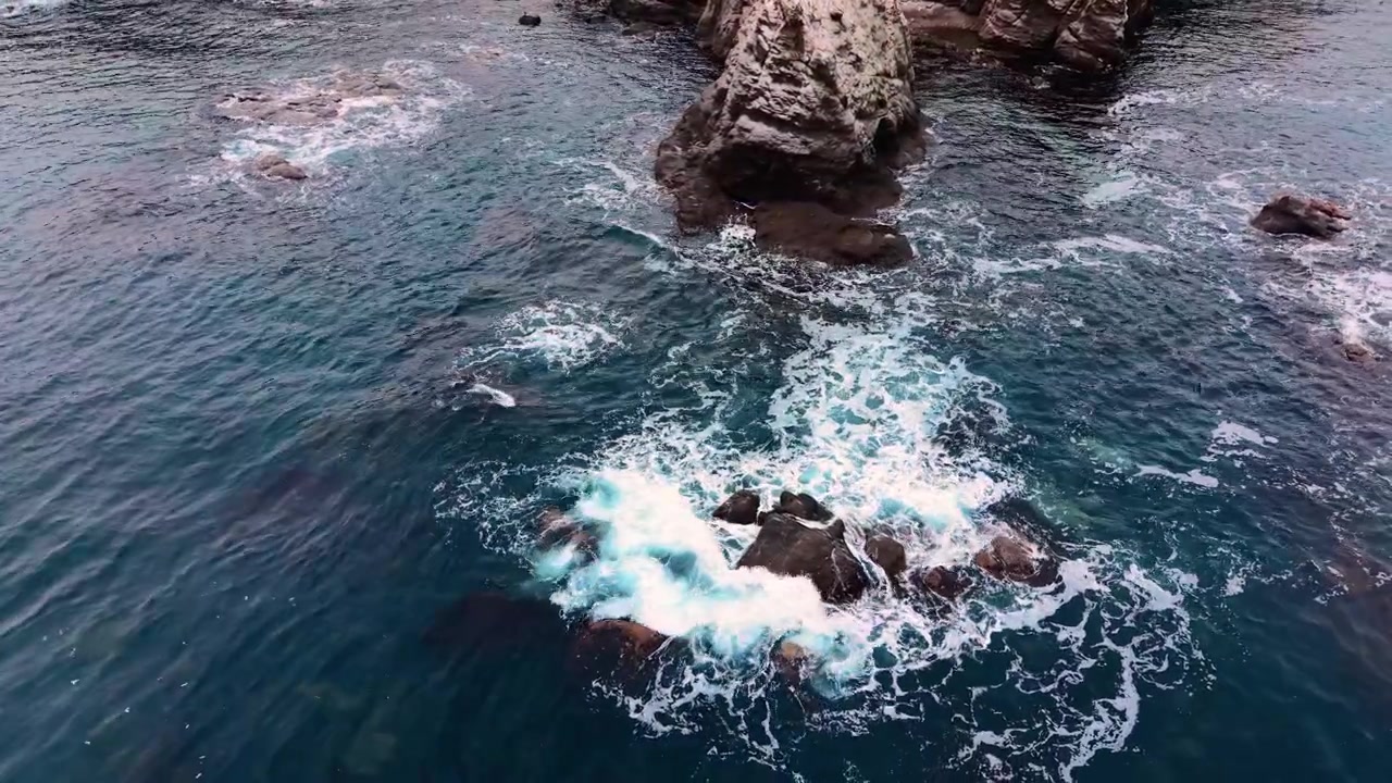 Prospettiva dall'alto di schiuma bianca e acqua turchese agitata attorno a massi di pietra. Onde oceaniche che si infrangono contro grandi rocce nel mare.