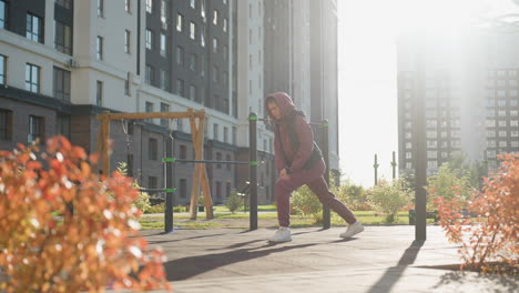 training partner in hoodie and quilted vest lunges forward under vibrant sunset light in urban park environment with fitness bars and walkers passing by warming up muscles for dynamic workout session
