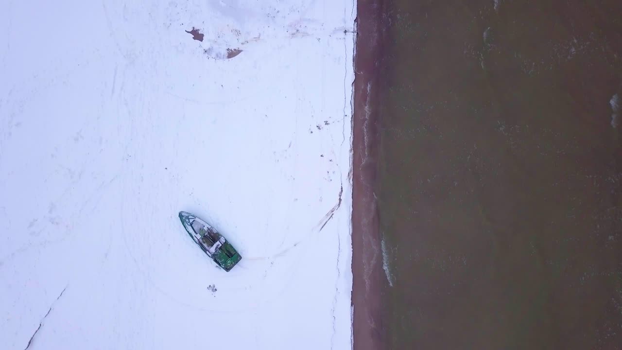 vista aérea de pájaro de la costa del mar báltico en un día de invierno nublado con un barco de pescadores costero verde, playa con arena blanca cubierta de nieve, erosión costera, gran tiro de drones moviéndose hacia atrás