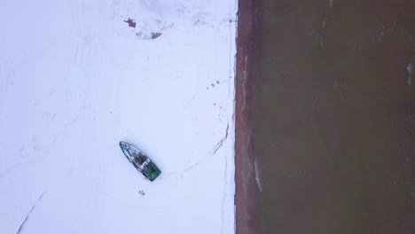 Aerial-birdseye-view-of-Baltic-sea-coast-on-a-overcast-winter-day-with-green-coastal-fisherman-boat,-beach-with-white-sand-covered-by-snow,-coastal-erosion,-wide-drone-shot-moving-backward