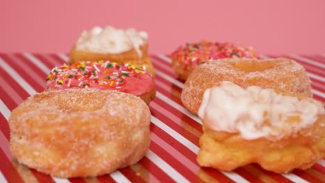 Delicious-pink-and-white-donuts-with-cream-frosting,-sugar-and-sprinkles-on-a-candy-cane-and-pink-backdrop---camera-shifting-focus-as-it-slide-in