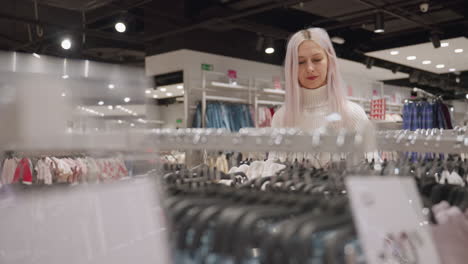 affectionate shopper walks to cloth rack and removes fleece pullover to admire fabric texture and stitch details with rose gold manicure under ambient boutique lighting beside moving shoppers