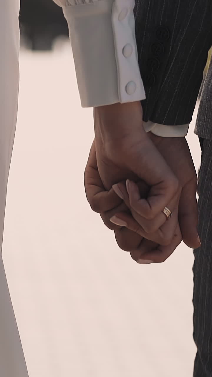 Bride in vintage dress and groom walk joining hands with interlaced fingers at wedding across park closeup slow motion. Newlywed couple in city