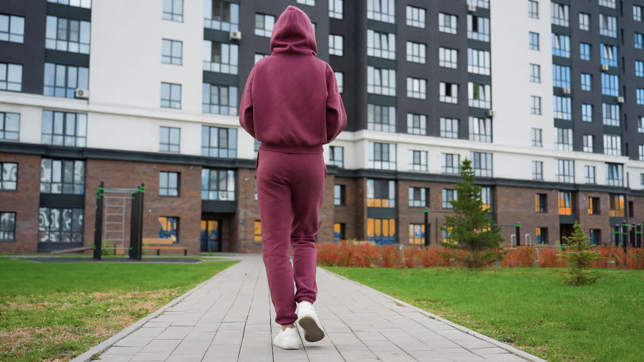 Back view of composed athlete holding water bottle walking toward gym building past blooming spring flowers along paved urban path under modern highrise facade on sunny spring morning