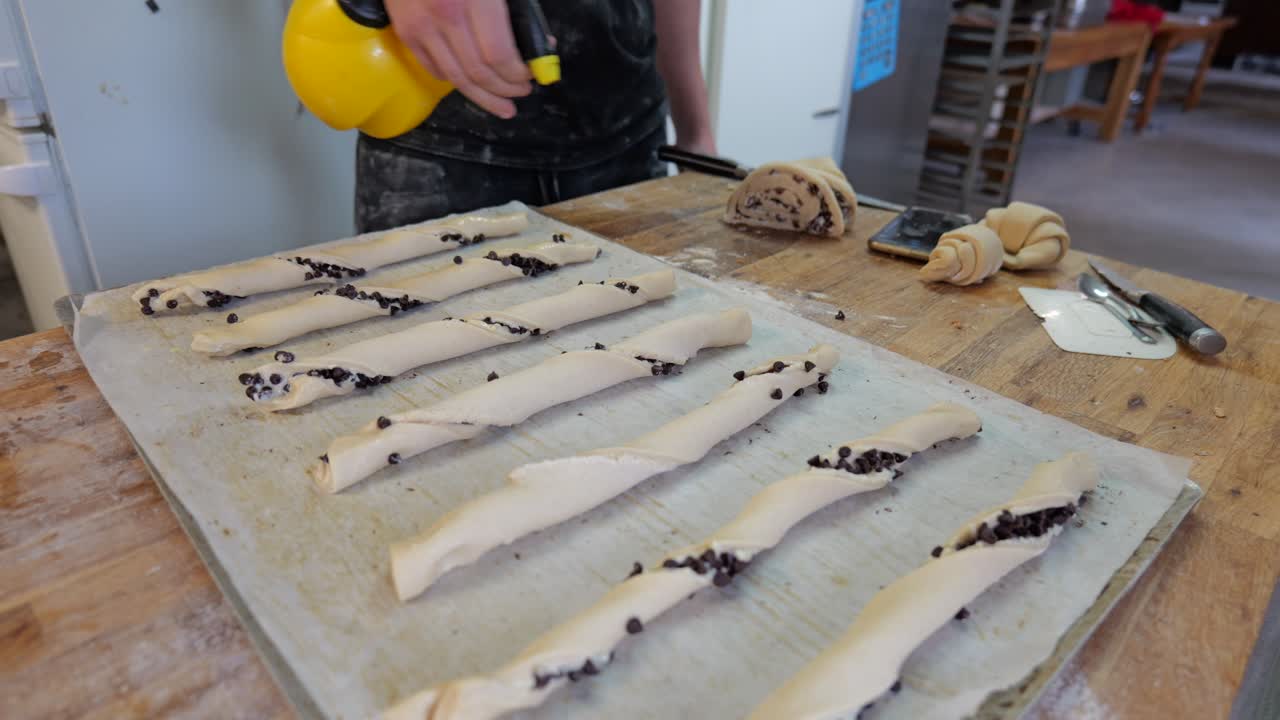 Baker sprays fat onto twisted chocolate pastries arranged on a baking tray
