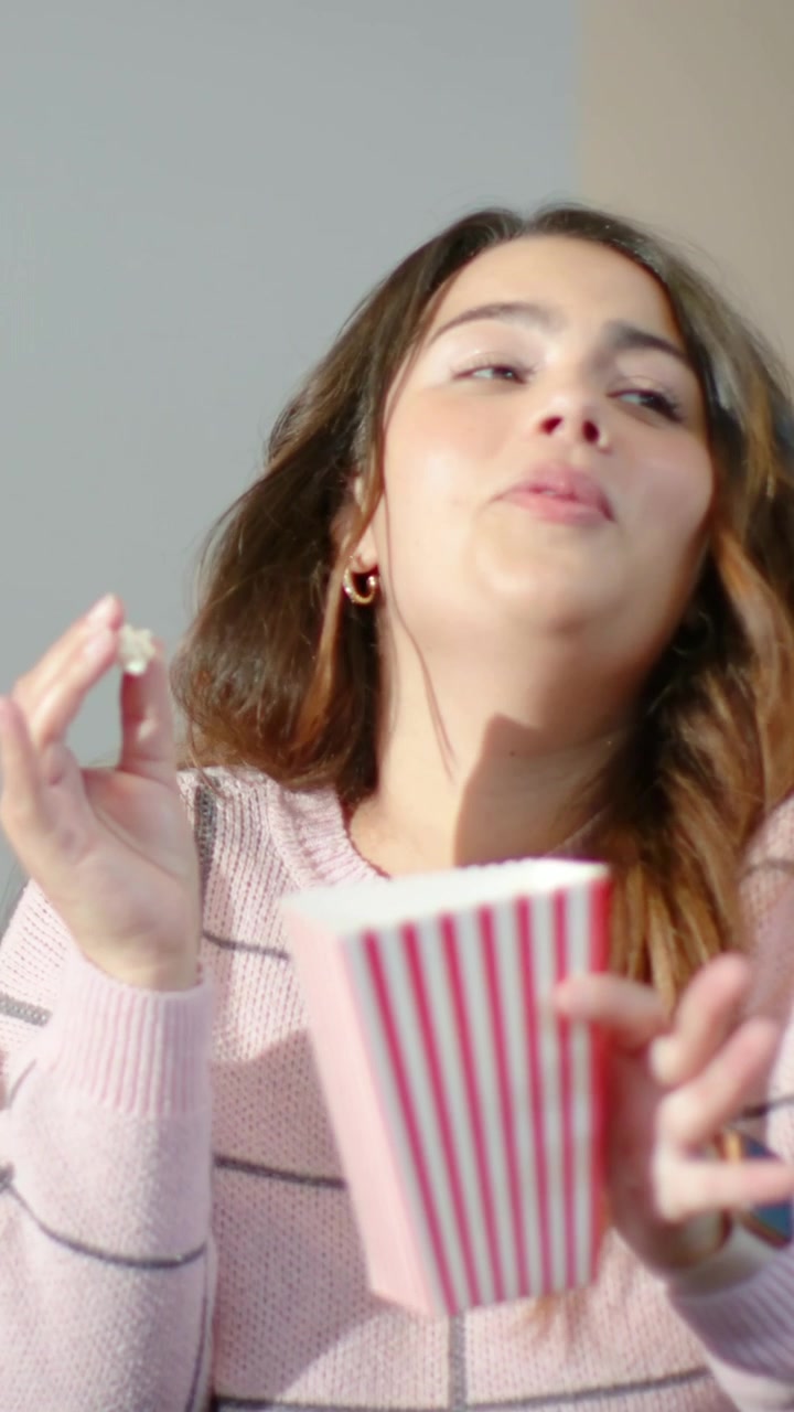Video verticale: Donna sorridente che prende e assaggia popcorn da una vaschetta a strisce in un loft, con indosso un maglione rosa.