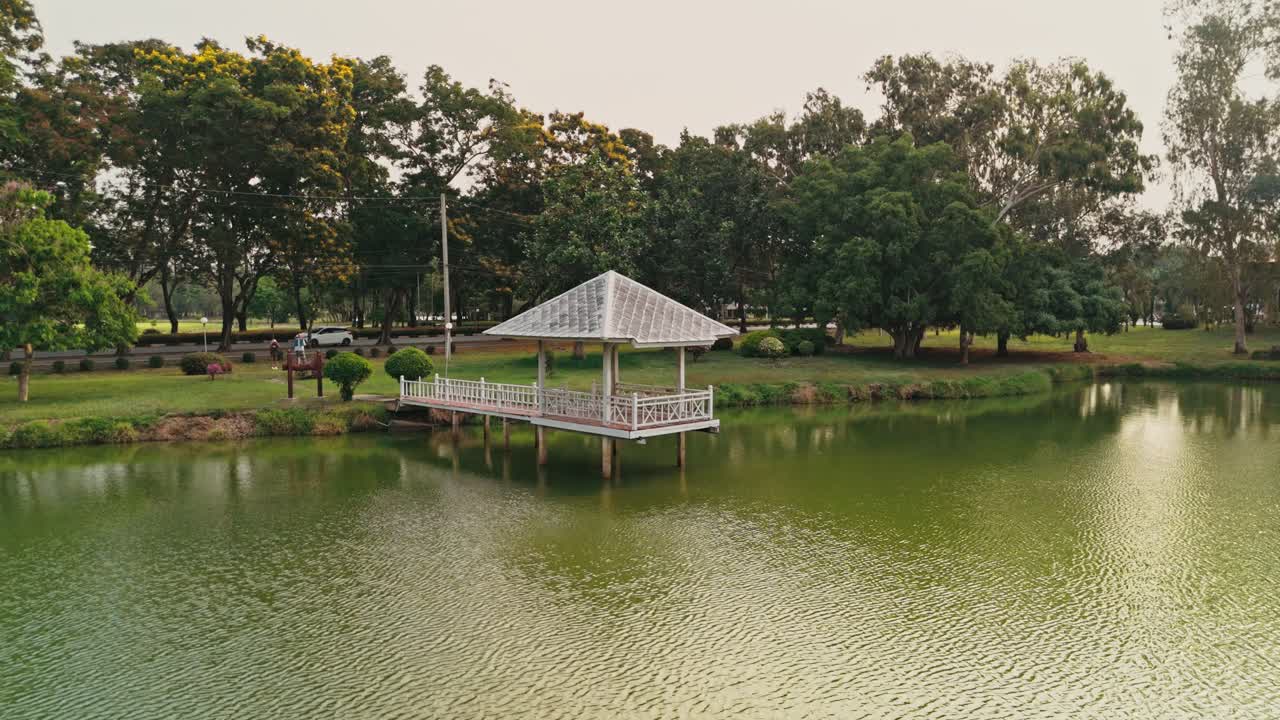 Et naturskønt luftfoto viser en smuk hvid pavillon, der strækker sig ud over en rolig grøn sø. Frodig tropisk vegetation omkranser den grønne bred, mens biler kører langs en stille vej i baggrunden i Thailand