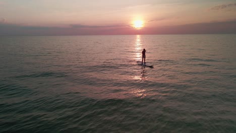 Vuelo-Aéreo-Lejos-Del-Hombre-Stand-Up-Paddle-Boarding-En-El-Océano-Al-Atardecer-O-Al-Amanecer