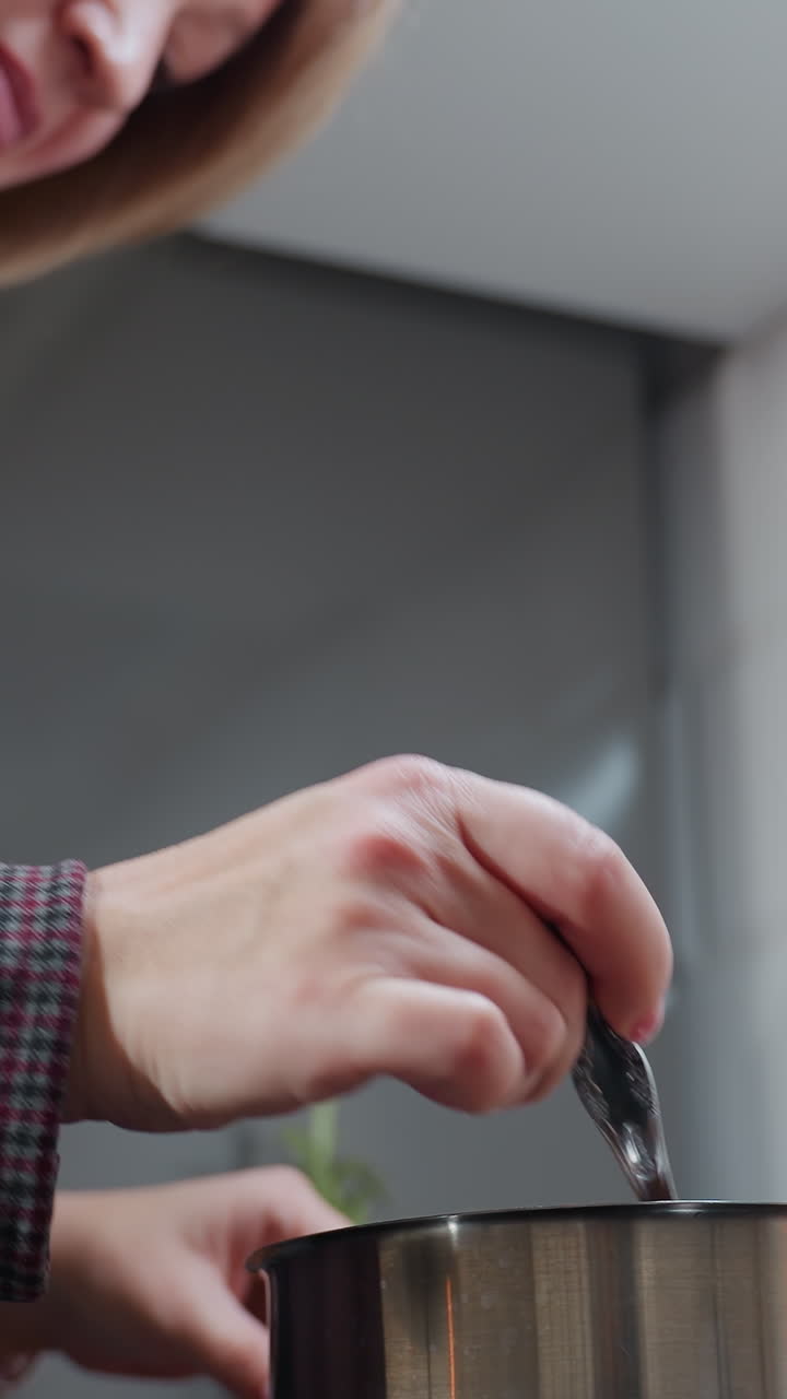 primer plano de una mujer agitando sopa en una olla de acero inoxidable en la estufa, con ropa de manga larga a cuadros, movimientos suaves de las manos, involucrada en el proceso de cocción, fondo de cocina de azulejos blancos