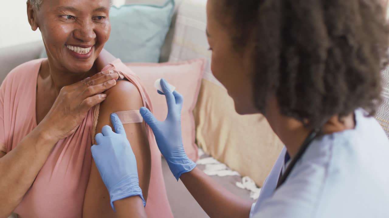 Une médecin afro-américaine posant un pansement sur l'épaule d'une femme âgée afro-américaine à la maison.