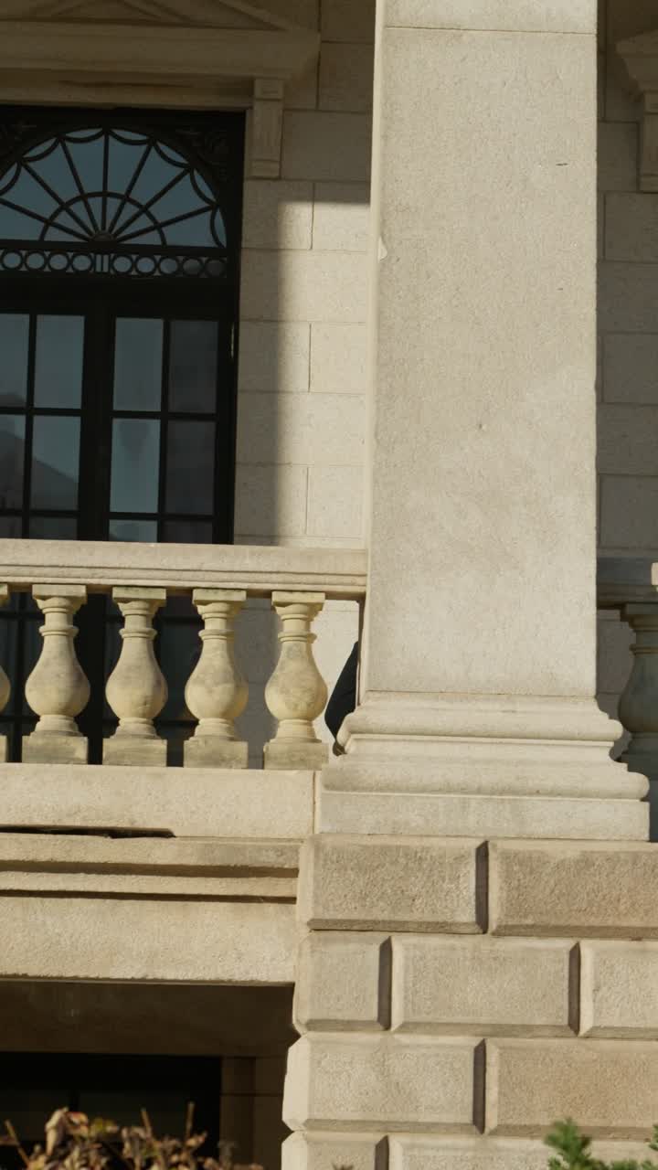 Stylish young woman in dark outfit and glasses walks past stone balustrade on sunlit neoclassical balcony of Seokjojeon palace in Seoul - slow motion, dolly tracking
