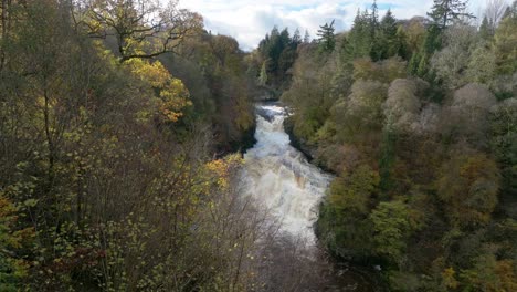 Stationary-drone-footage-of-a-cascading-waterfall-in-a-gorge-surrounded-by-colourful-trees-in-autumn