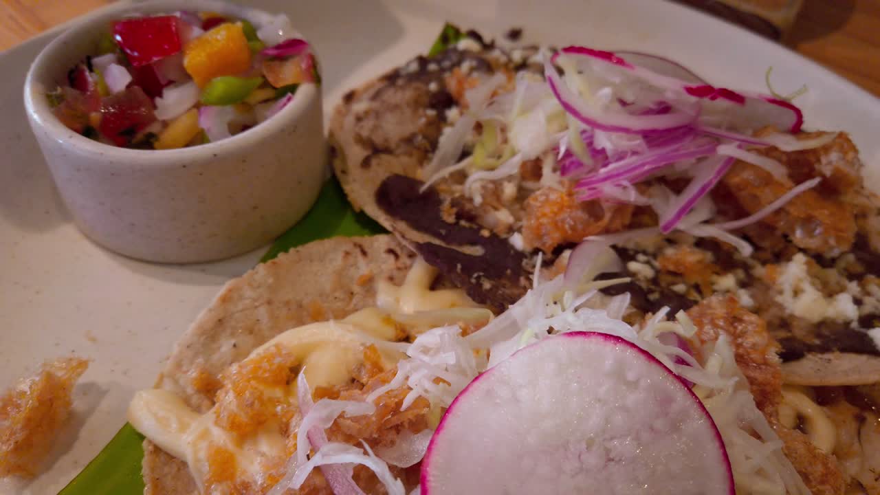 Close-up of a savory Oaxacan tlayuda layered with black beans, cheese, fried pork skin, radish, pickled onions and lettuce, served with a side of spicy salsa