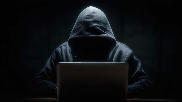 Una figura misteriosa con una felpa con cappuccio, impegnata in attività sospette davanti a un laptop, circondata dall'oscurità e da ombre enigmatiche.
