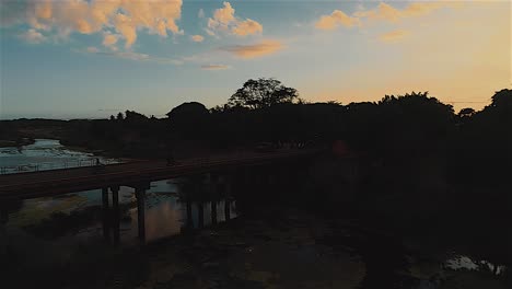 Flying-over-Nizao-river-with-cars-and-motorcycle-crossing-bridge-in-San-Cristobal,-Dominican-Republic