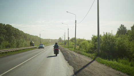 back view of two friends riding a power bike slowly down a road lined with street lamps, with a car coming in opposite direction, with trees and a signpost visible in the background