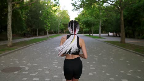 slow motion rear view of european woman with black and white dreadlocks running by local, green park in the city. she is