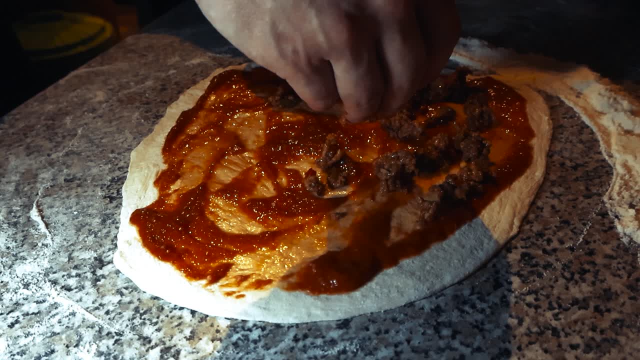 Slow motion low angle view around home cook hand adding minced meat topping over a raw pizza base with tomato sauce in closeup. Applying ingredients on fresh homemade dough on dimly lit kitchen table