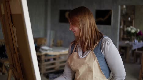 portrait of caucasian woman painting in arts studio using smear and oil paints. long haired artist in beige apron, smiling while process, looking to the camera. slow motion