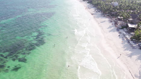 Toma-Aérea-De-Olas-En-Una-Playa-De-Arena-Blanca-Rodeada-De-Cabañas-Y-Cabañas-Con-Palmeras-En-Un-Océano-Azul-Cristalino-En-Tulum,-México.