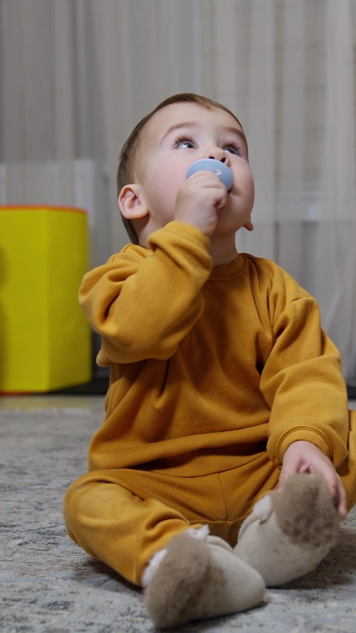 Calm cute baby boy sits on the floor holding a pacifier at his mouth. Kid looks around the room ignoring two bright pyramids beside. Vertical video